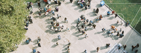 An aerial view of a scattered group of people standing on a pavement with a tree on the left and grass on the right. There is also a Verian triangle graphic shape overlaying the right-hand side.
