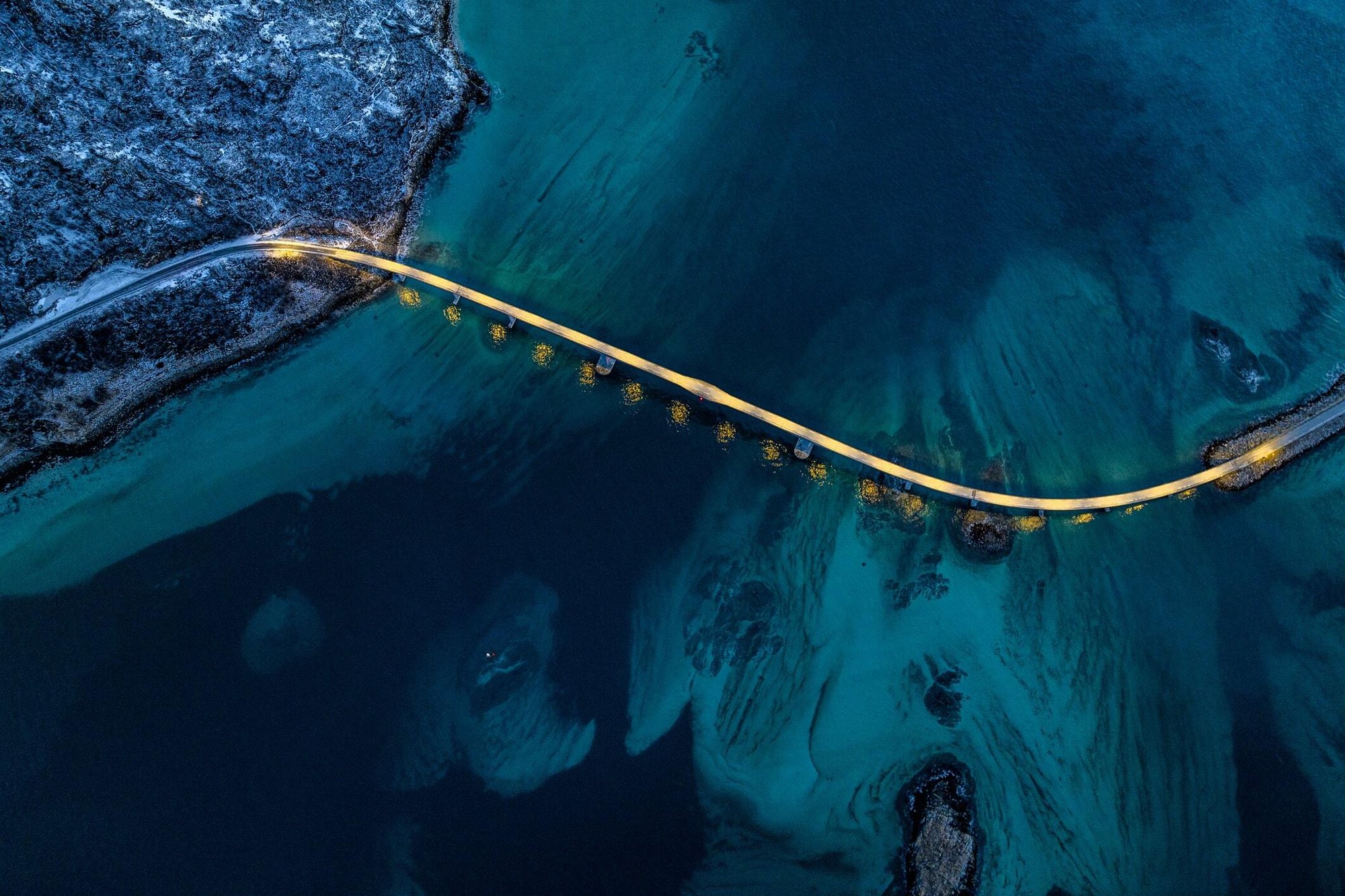An aerial, nighttime view of a narrow, illuminated road bridge curving across a turquoise and deep blue ocean channel, connecting a snow-dusted, rocky coastline to another piece of land.