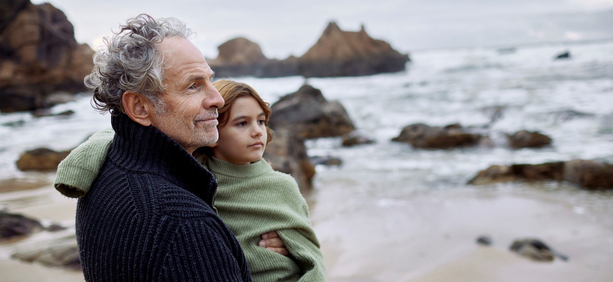 Person holding a child on a rocky beach overlooking the sea.
