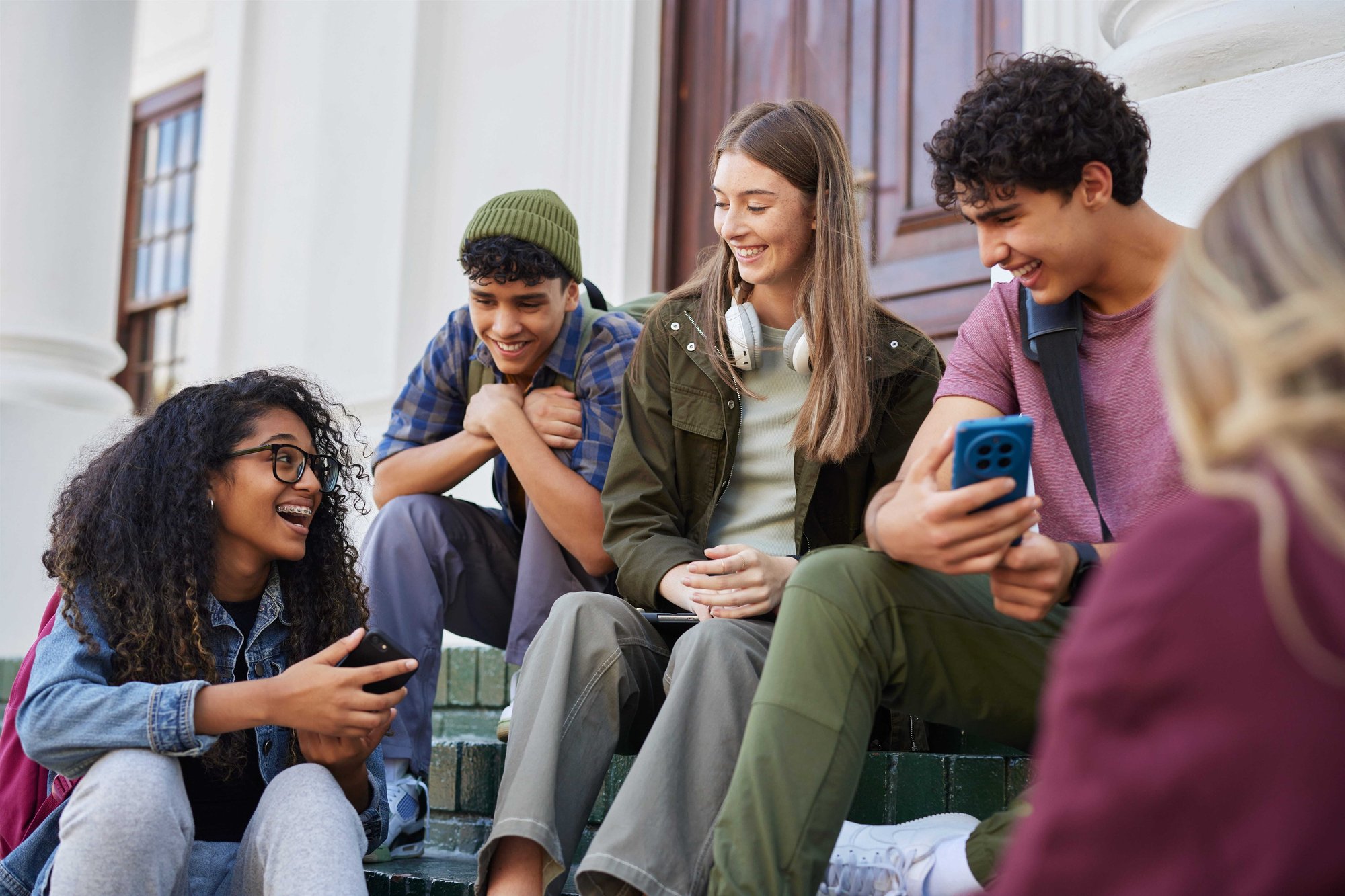 A diverse group of five smiling young adults sitting on outdoor stone steps, engaged in conversation and looking at their smartphones.