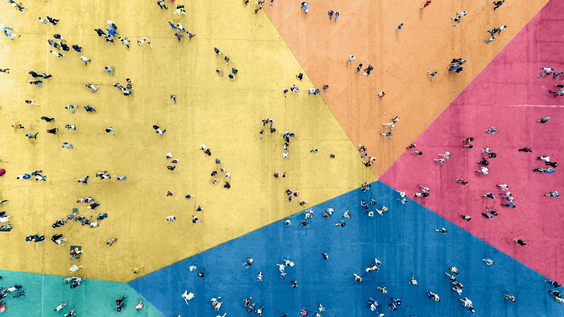 Abgebildet ist eine große Gruppe von Leuten, die auf einem großen Platz unterwegs sind. Die Aufnahme ist aus der Vogelperspektive entstanden und der Platz, auf dem sich die Leute aufhalten, ist in fünf bunte Zonen eingeteilt, die jeweils eine andere Farbe haben.