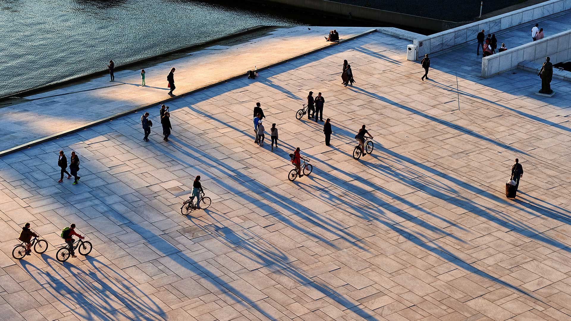 Ein großer Platz, welcher zudem am Wasser liegt. Auf dem Platz sind vereinzelt Leute zu sehen, die entweder zu fuß oder auf dem Fahrrad unterwegs sind.