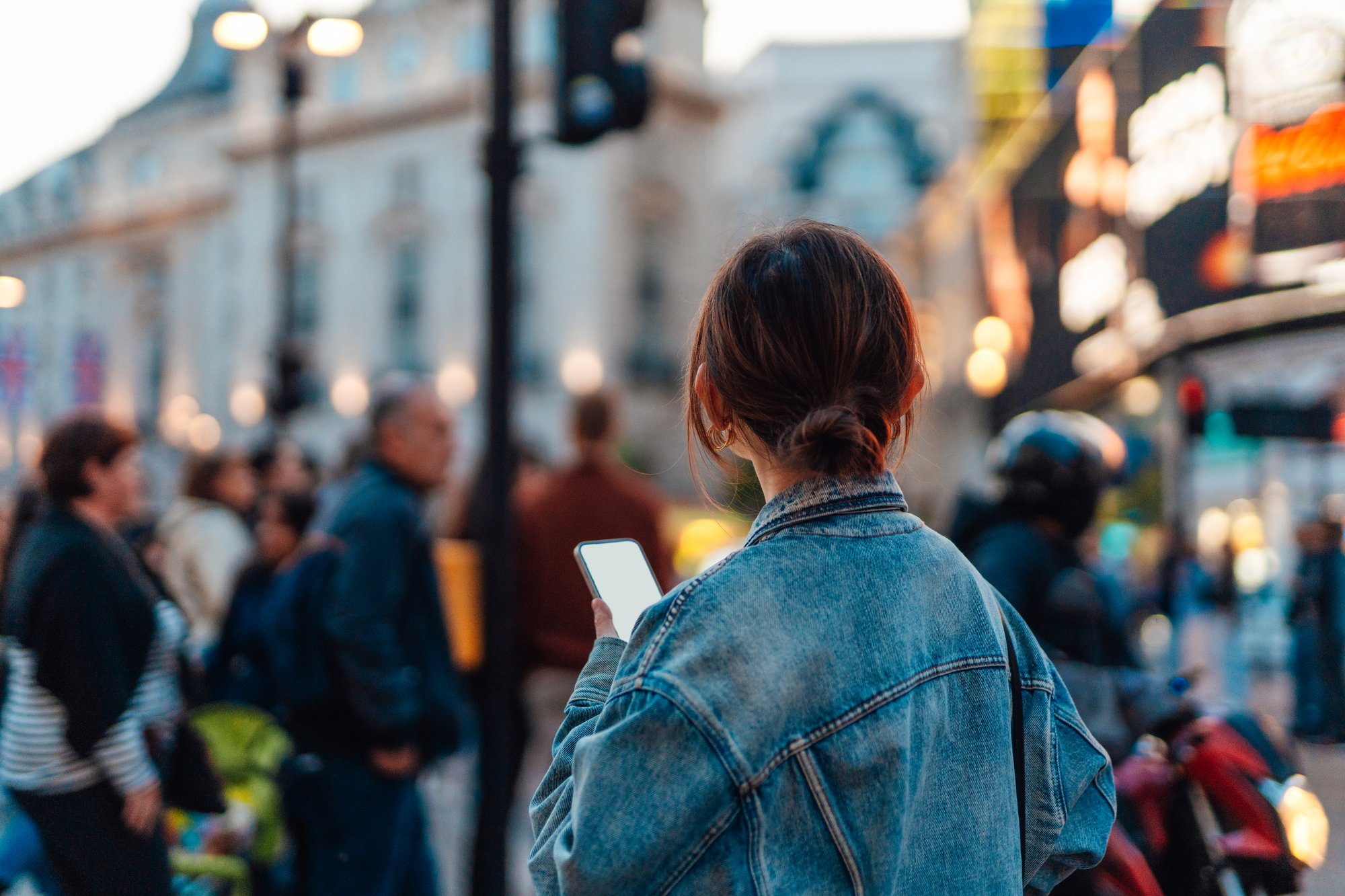 Auf dem Bild ist eine Frau abgebildet, die uns den Rücken zukehrt und ein Handy in der Hand hält. Sie schaut nach vorne in eine Menschenmenge, die für uns unscharf dargestellt ist.