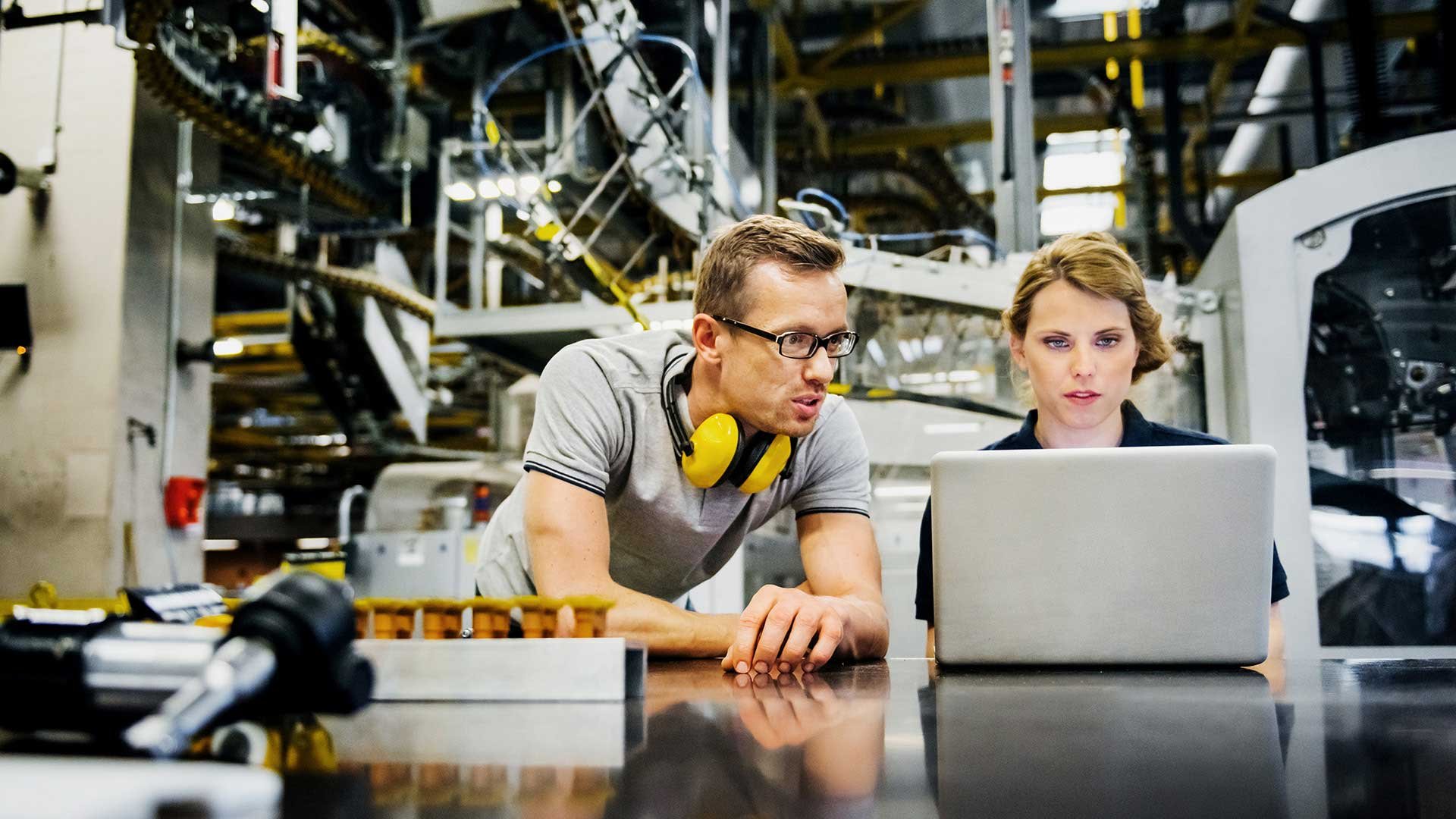 Auf dem Bild sieht man einen Mann und eine Frau in einer Fabrik oder Produktionsanlage. Die Frau sitzt vor einem Laptop und der Mann schaut auf den Bildschirm des Laptops. Beide scheinen miteinander zu sprechen.