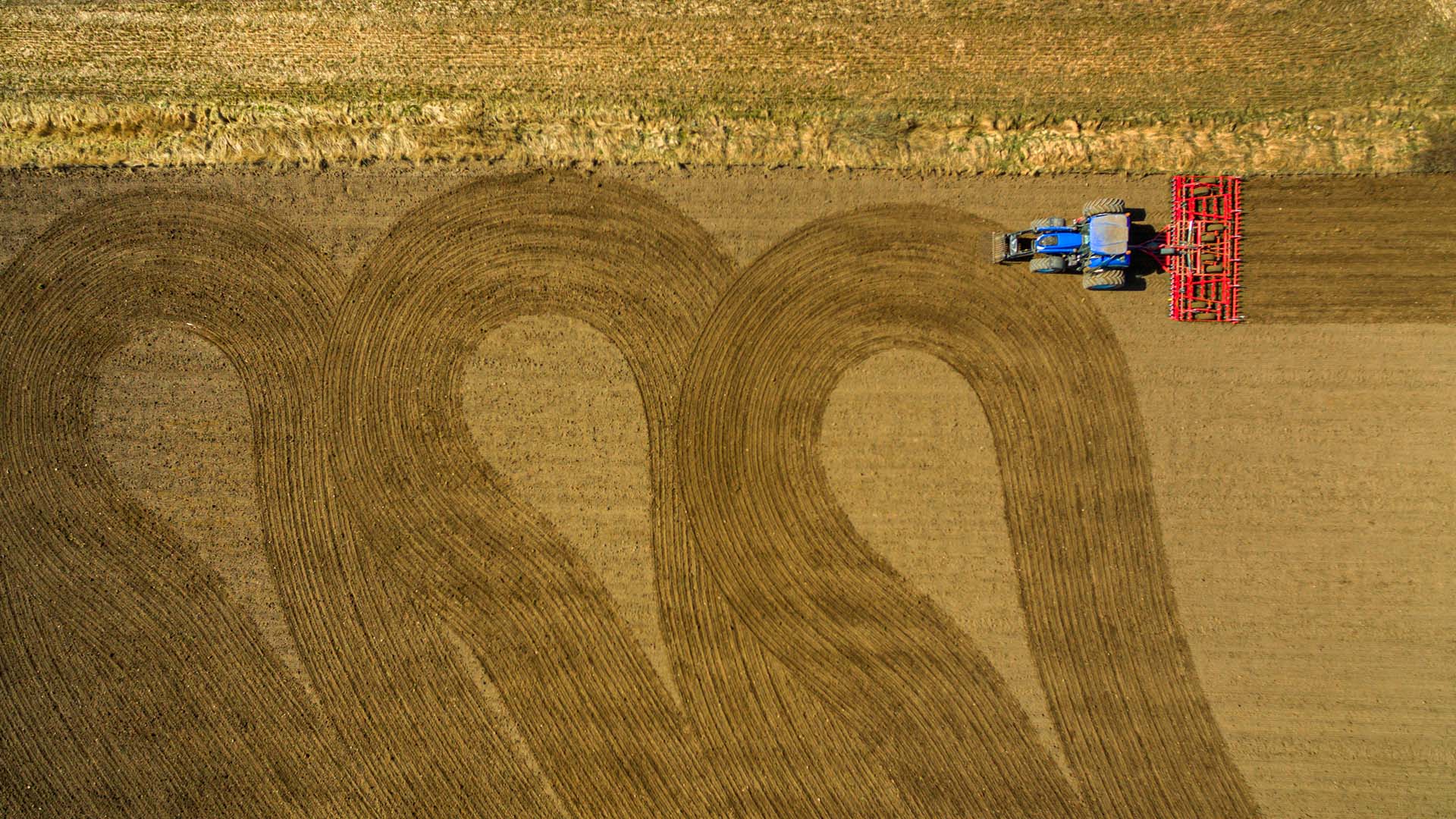 Flygbild uppifrån av en åker som bearbetas: en blå traktor med rött jordbruksredskap kör längs kanten och lämnar breda, böjda plogfåror i den bruna jorden.