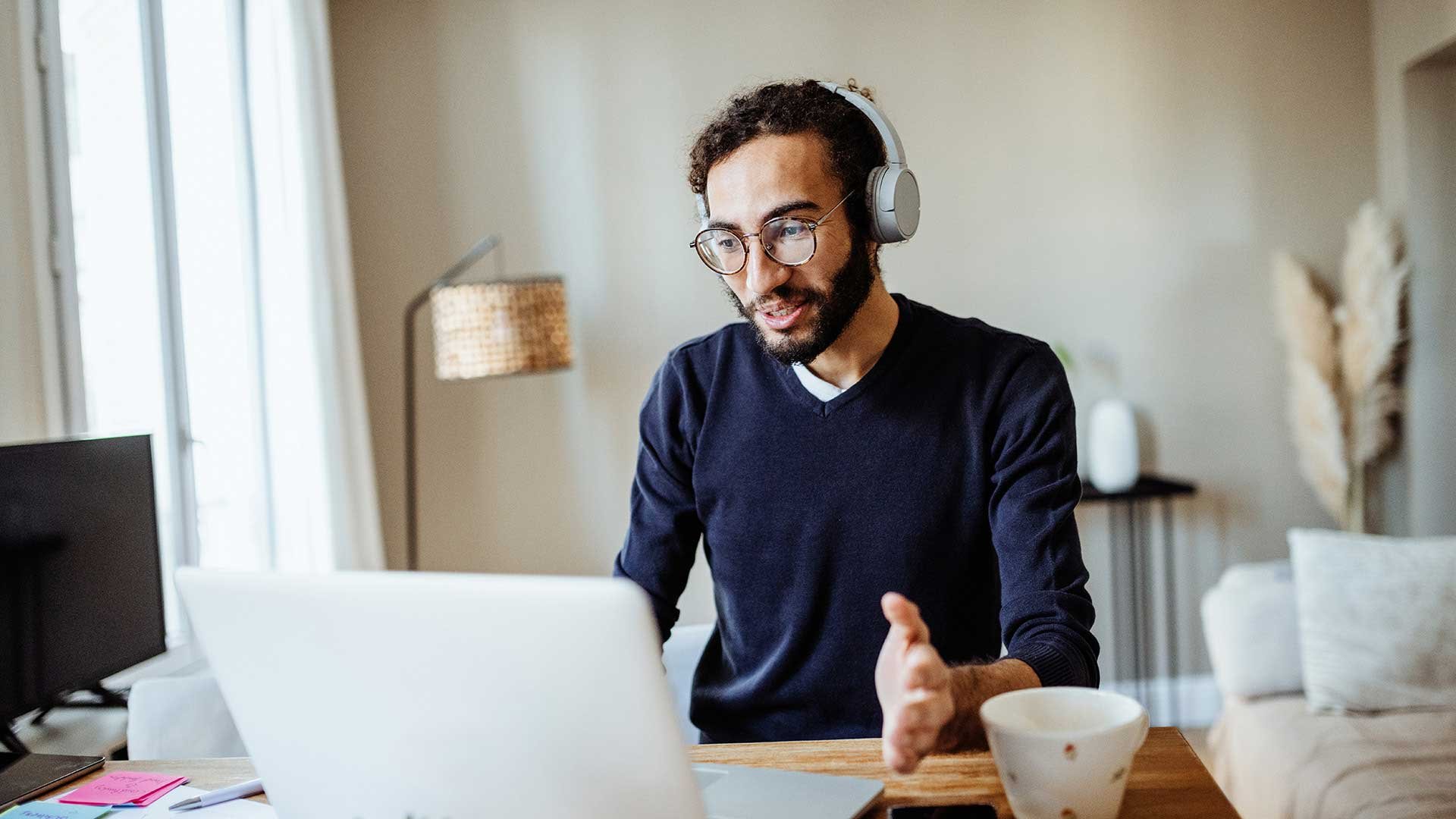 En man arbetar hemifrån. Han har hörlurar på och pratar med någon i ett onlinemöte, datorskärmen syns bakifrån. Mannen har glasögon, mörkt skägg och hår.