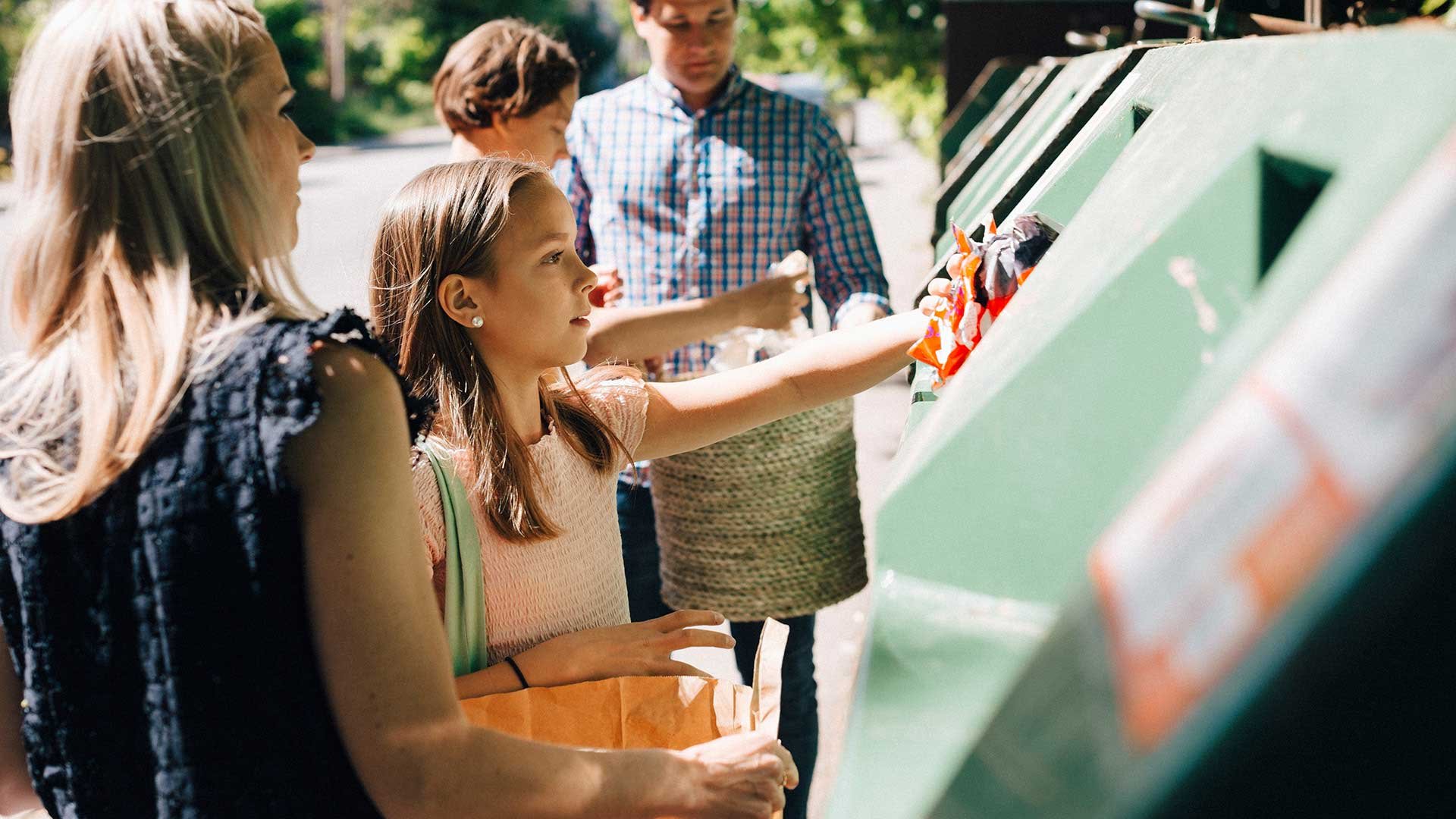 Barn och vuxna vid returstationens gröna behållare, återvinner papper och plast. Solen lyser, det är sommar.