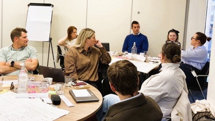 Participants gathered around tables in a workshop session, discussing ideas and reviewing notes and materials together.