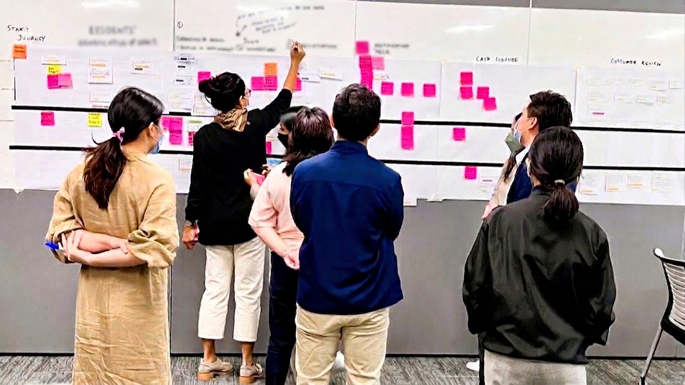 Workshop participants review a service journey map on a wall, using sticky notes and process diagrams to discuss and refine workflows.